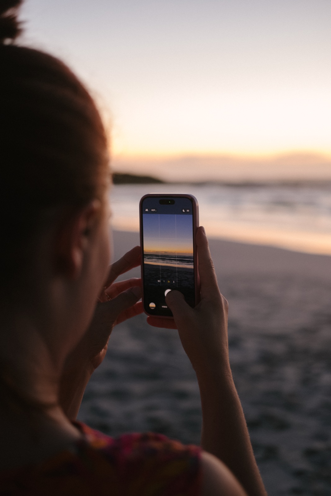 Over-the-shoulder view of Jane framing a beach sunset with her iPhone